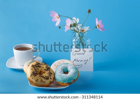 Concept breakfast. Donuts on saucer with cup of tea, flowers  and note "Good morning"