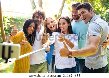 Happy friends making selfie photo on smartphone while showing thumbs up outdoors