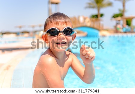 Happy grinning young boy at a swimming pool sitting on the edge wearing his goggles and costume as he enjoys the summer sun on vacation