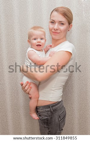 Mother holding and carrying her baby at home. Baby like mother. Family look in white t-shirts. 