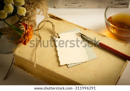 Old book with decorative greeting card, nib pen and cup tea on white wooden boards 