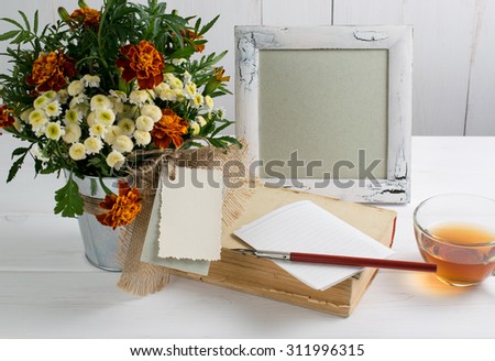 Tagetes flower bouquet with decorative greeting card, photo frame and old book with cup tea and nib pen on white wooden boards in shabby chic style