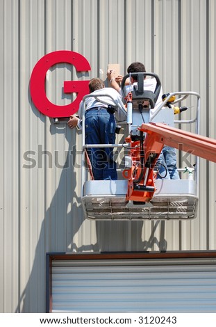 A pair of workmen install a sign on the side of an industrial building using a lift.