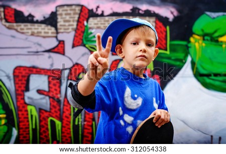 Young boy making a V-sign gesture for success and victory, or peace, as he stands in front of a graffiti covered wall holding his skateboard