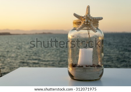 decorative glass jar with sand, a candle and a starfish on a background of calm sea and the evening sky is on the table. The perfect setting for a romantic dinner on the beach at sunset