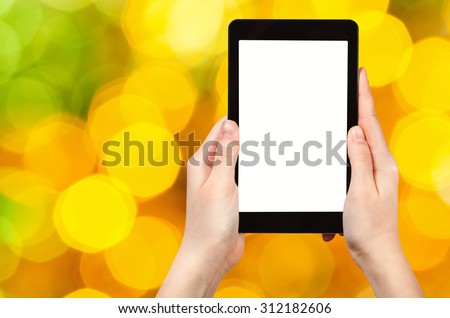 christmas party concept - hand with tablet pc with cut out screen on background from big yellow and green shimmering Christmas lights of electric garlands on Xmas tree