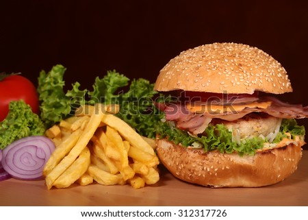 One big tasty appetizing fresh burger of green lettuce red tomato cheese bacon slice meat cutlet violet oinion white bread bun with sesame seeds and chips on wooden table closeup, horizontal picture