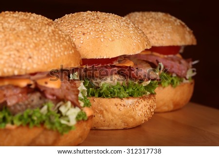 Three appetizing big delicious fresh burgers with green lettuce red tomato cheese cabbage bacon slice meat cutlet and white bread bun with sesame seeds on black background closeup, horizontal picture