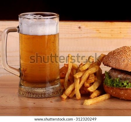 Big fresh tasty burger of green lettuce meat cutlet cheese tomato and white bread bun with sesame seeds near chips and glass of light beer on octoberfest holiday on wooden table, square picture