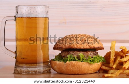 Big fresh tasty burger of green lettuce meat cutlet cheese tomato and white bread bun with sesame seeds near chips and glass of light beer on octoberfest holiday, horizontal picture