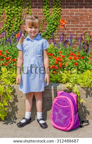 Preschool girl with braids dressed in school uniform gingham blue dress and black classic mary jane shoes standing near the school wall with her backpack. Back to school.
