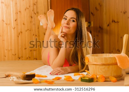 Woman with bucket ladle and petals relaxing in finnish sauna. Attractive girl in bikini resting. Spa wellbeing pleasure.