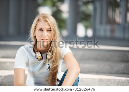 Beautiful young woman with vintage music headphones around her neck, sitting against urban city background and looking straight.