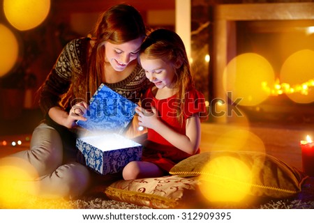 Young mother and her little daughter opening a magical Christmas gift by a Christmas tree in cozy living room in winter