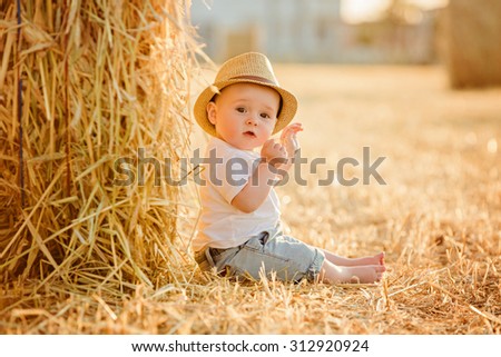 Little adorable baby boy with big brown eyes in a hat sits in a field near haystacks at sunset in the summer and looks serious eyes