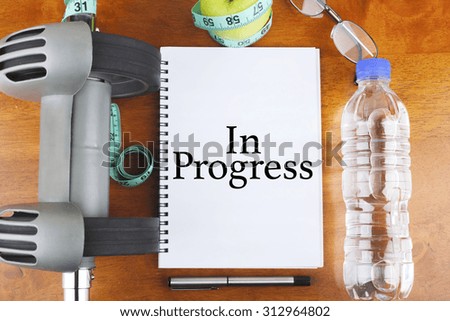 "In Progress" text on notebook with delicious green apple, measure tape, spectacle, a bottle of mineral water, and bodybuilding tools on wooden background - healthy, exercise and diet concept