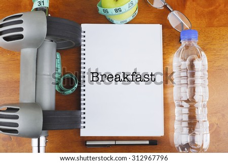 "Breakfast" text on notebook with delicious green apple, measure tape, spectacle, a bottle of mineral water, and bodybuilding tools on wooden background - healthy, exercise and diet concept