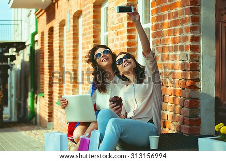 Two beautiful young brunette student sitting on the steps on the street with a backpack and make self-photo. Two friends or sisters with laptop after shopping resting on the steps and make a photo.
