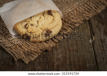 Chewy chocolate chip cookie, fresh from the oven. Sat on a hessian and wood surface. Cookie is individually wrapped in grease proof paper Royalty-Free Stock Photo #313191734