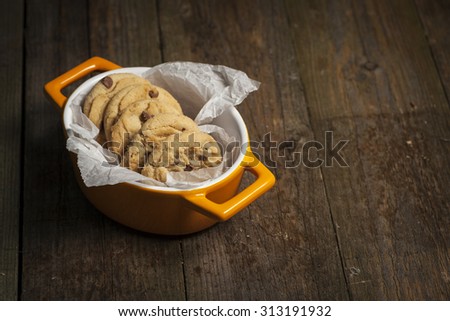 Chewy chocolate chip cookies, fresh from the oven in a small orange dish lined with grease proof paper. Sat on an old wood surface. Royalty-Free Stock Photo #313191932