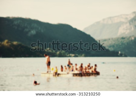 Blurred photo of Annecy lake (France). Adults and kids relax on rubber floating raft  at the Annecy lake surrounded by beautiful mountains. Toned photo.