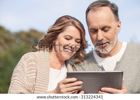 Pretty mature man and woman are holding a laptop and looking at it with joy. They are embracing and smiling. The family is resting in park
