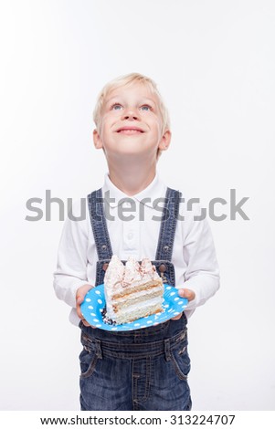 Pretty male child is holding a plate with large tasty piece of cake. He is standing and smiling. The schoolboy is looking up thankfully. Isolated on background