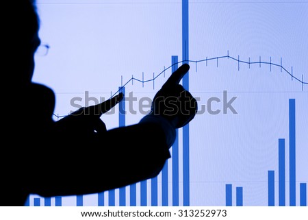 Silhouette of a man pointing with both hands to a rising graph on an LCD display. Focus is on the LCD display.