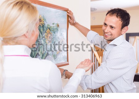 Young man and blonde woman hanging art picture in frame. Focus on man