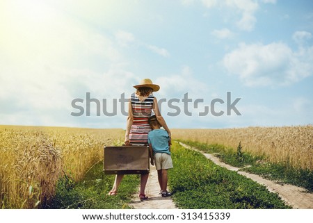 Picture of woman and little boy standing in middle of country road. Mother and son travelling together with old suitcase on summer sky outdoor background.