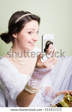 Picture of happy young girl in white lace dress making selfie with smartphone. Pretty woman with pearls smiling on blurred olivine indoor background.