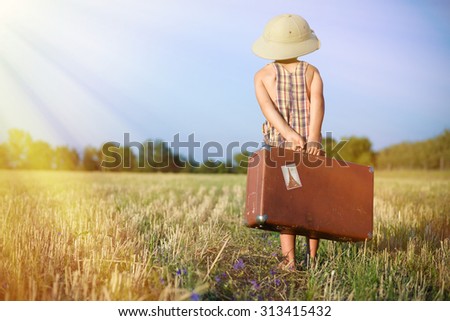 Picture of little boy wearing pith helmet carrying old suitcase in countryside. Backview of kid in plaid romper walking away on sunny flare background.