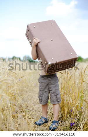 Picture of little boy lifting up big old suitcase in wheat field. Full length kid under heavy brown valize over blue sky background