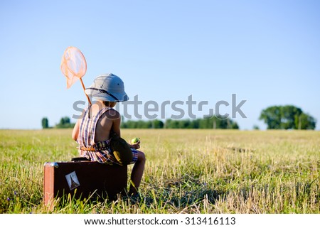 Picture of sweet small boy sitting on old brown suitcase eating green apple. Kid has ring net and flask over blue sky sunny outdoors background