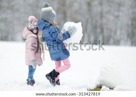 Two funny adorable little sisters building a snowman together in beautiful winter park during snowfall