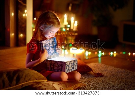 Adorable little girl opening a magical Christmas gift by a Christmas tree in cozy living room in winter