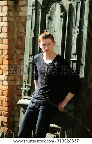 One cute red haired young fashionable unshaven stylish man in black jersey and jeans standing leaning on wooden green door outdoor sunny day on brick wall background, vertical picture