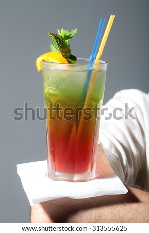 One beautiful alcoholic long cocktail red and green colors in drinking glass with straws mint leaf and yellow lemon slice standing on human hand on grey studio background, vertical picture