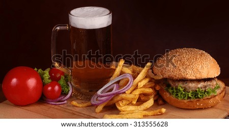 Big fresh tasty burger of green lettuce meat cutlet cheese tomato and white bread bun with sesame seeds near chips and glass of dark beer on octoberfest holiday on brown background, horizontal picture