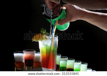 Colorful alcoholic shot cocktails in glasses and barman hand pouring from shaker into long beverage with straws mint and orange standing in row on black studio background, horizontal picture