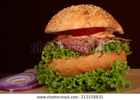 One big tasty appetizing fresh burger of green lettuce red tomato cheese bacon slice cabbage violet oinion and white bread bun with sesame seeds on black background closeup, horizontal picture