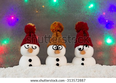 Three snowmen with knit caps sitting in the snow in front of a colorful background
