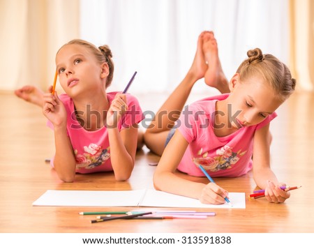 Young twins sisters are drawing on paper with colored pencils lying on the floor.