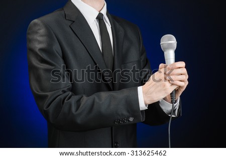 Business and speech topic: Man in black suit holding a microphone on a gray dark blue isolated background in studio