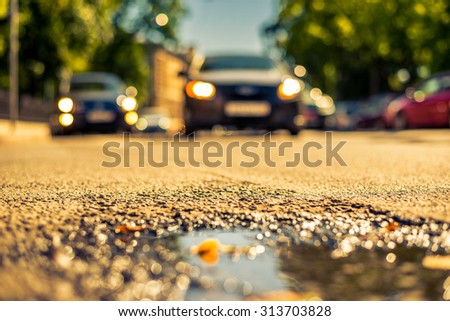Clear day in the big city, car approaching on a city street near the park. View from puddles on the pavement level, image in the yellow-blue toning
