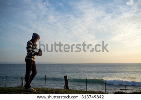 Young Woman Standing on the Grassy Landscape at the Seaside and Taking Pictures of the Beautiful Sea During Sunset.