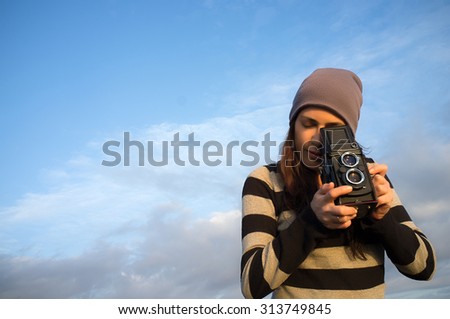Pretty young woman taking pictures using a vintage camera outdoors. Lifestyle portrait over blue sky with copy space at the left.