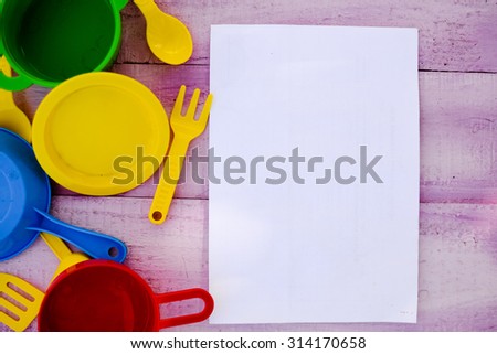 Picture of wooden tabletop with colorful toy dishware and blank sheet of paper. Plastic pates and cutlery on lilac rustic with copy space background.