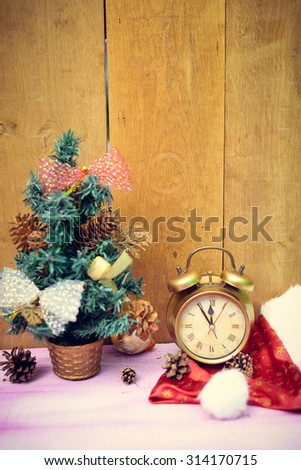 Picture of little christmas tree decorated with bows and old alarm clock. Artificial tree, pine cones and santa hat on wooden indoor background.