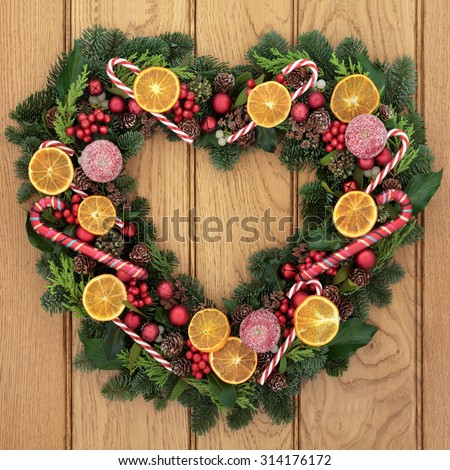Christmas heart shaped wreath with dried fruit, candy canes, bauble decorations, holly, mistletoe and greenery over oak front door background.